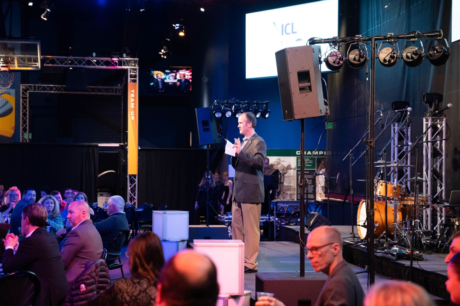 A speaker standing in front of a seated crowd and addresses the party.