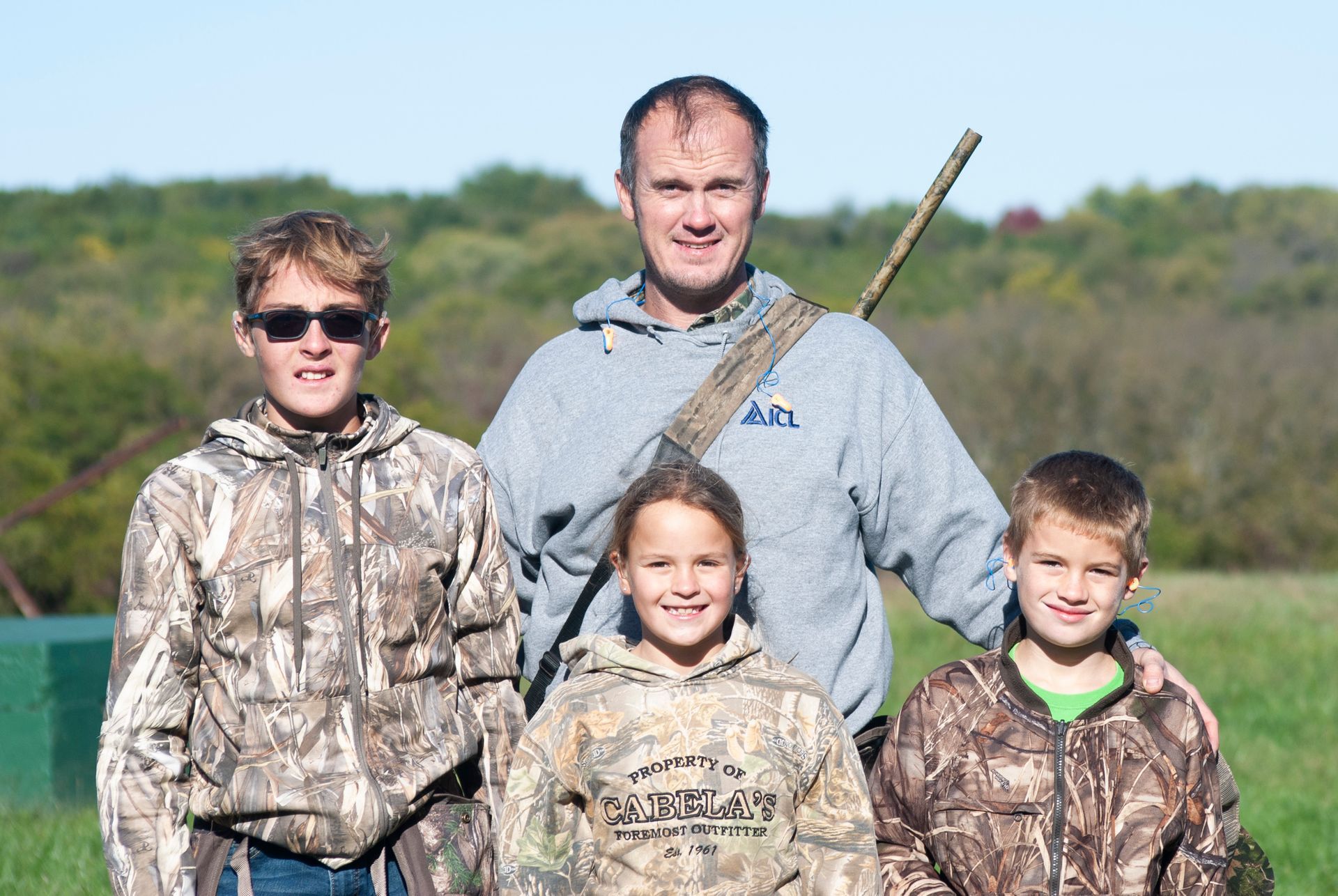 Three kids stand with their dad as they wear camouflage and the dad holds a gun on his back.