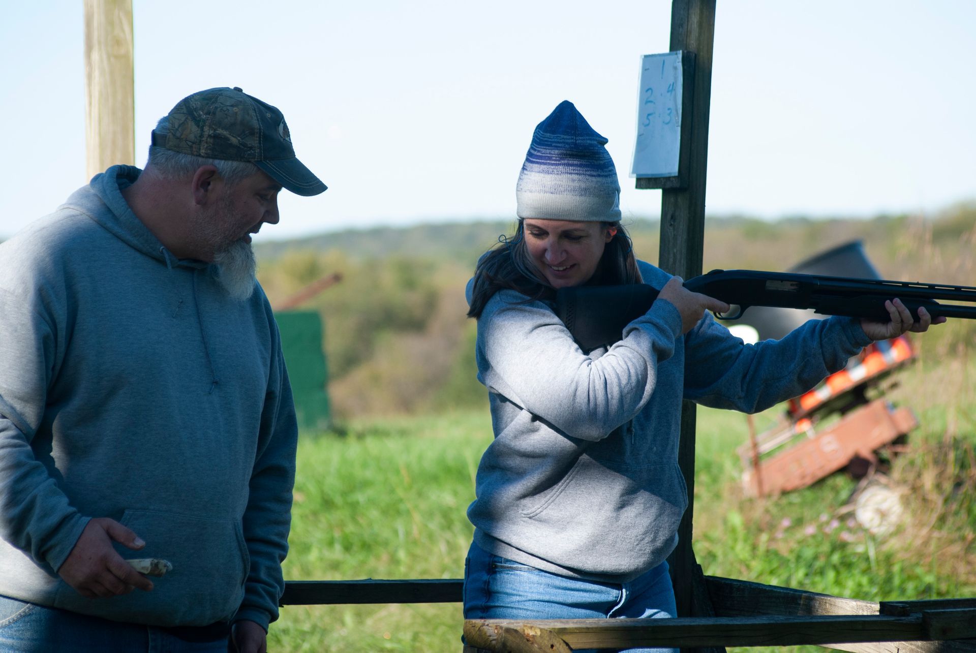 A woman is talking to a man as she holds a gun and gets ready to skeet shoot.