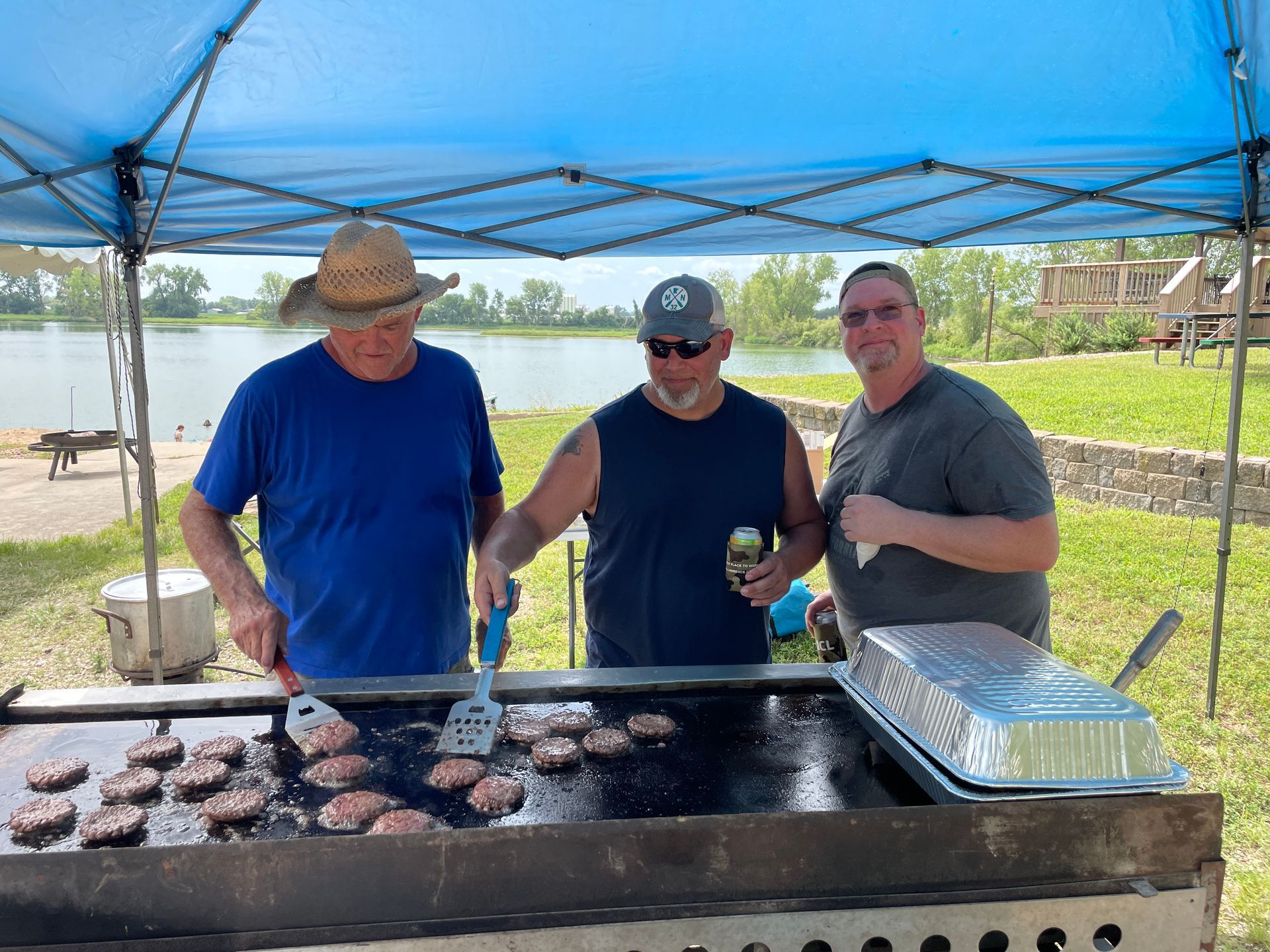 three icl employees are cooking hamburgers on a grill under a blue tent .
