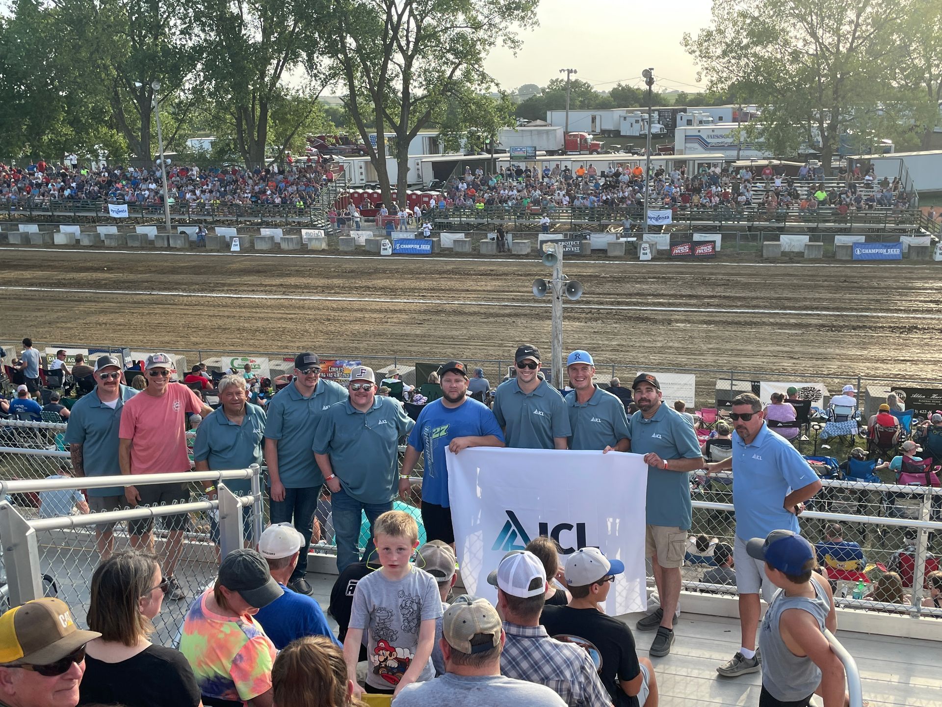 a group of icl employees are standing in front of a crowd at a race track holding a flag .