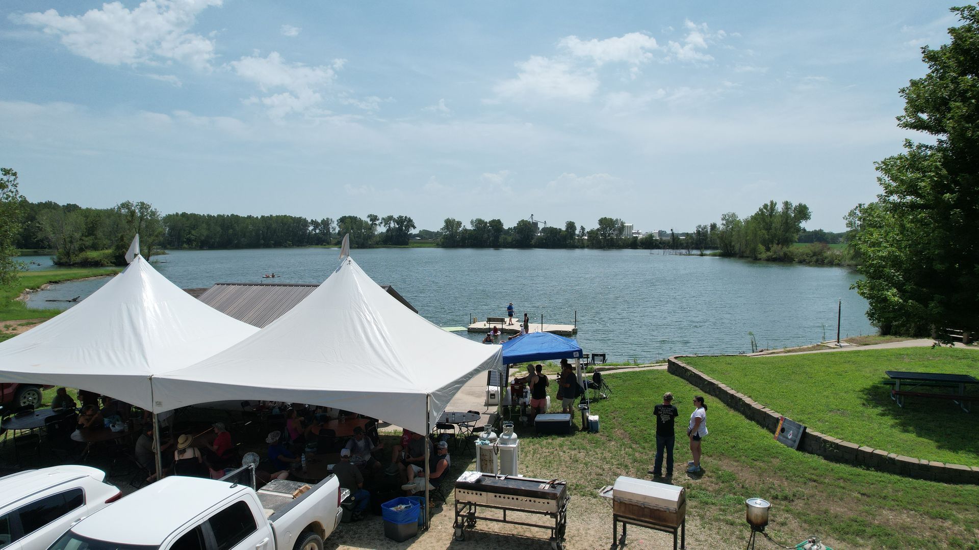 Two large white tents are erected near a lake.