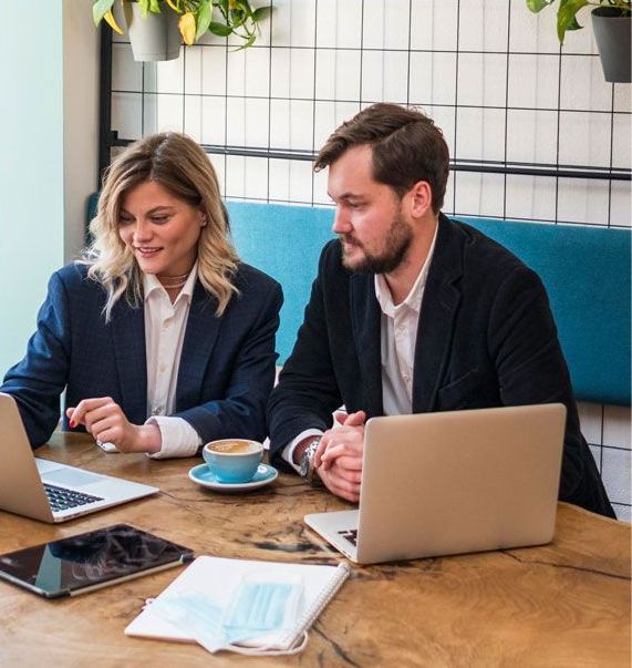 Two business professionals, a man and a woman, looking at laptop screens while sitting at a table with coffee, tablets, and a mask.