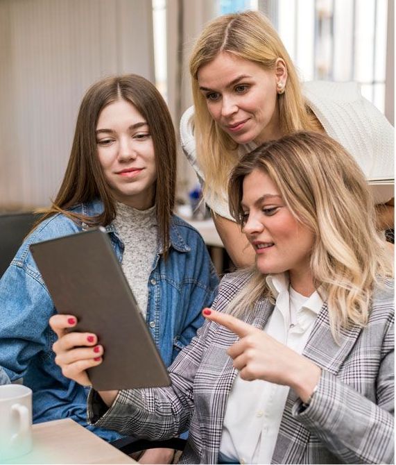 Three women looking at a tablet, one pointing. They're in an office setting, discussing something on the screen.