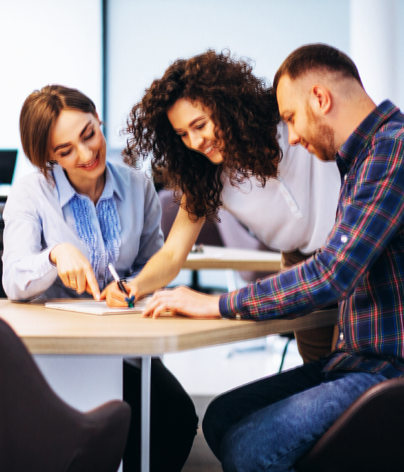 Three colleagues collaborate around a table, reviewing paperwork. A woman points to a document, while the others observe and smile in an office setting.