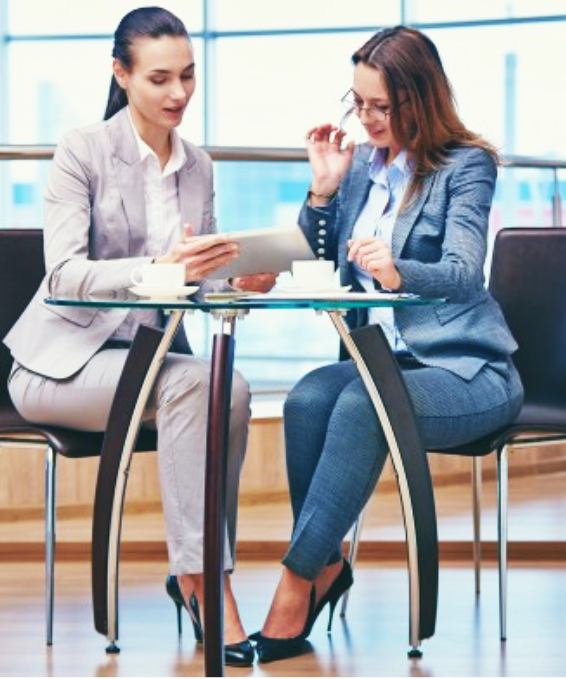 Two women in business suits seated at a table, reviewing a tablet, in a modern office setting.
