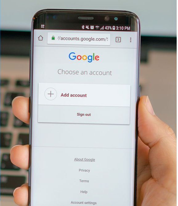 A hand holding a smartphone displaying a Google account login screen with the options to add an account or sign out.