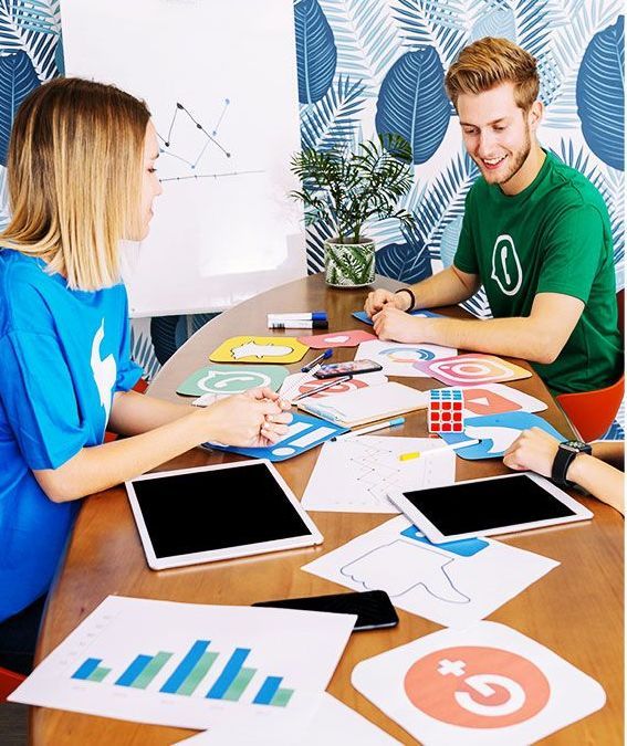Two people, a woman in blue and a man in green, at a table with papers, tablets, and a Rubik's Cube. They are collaborating, likely on a project.