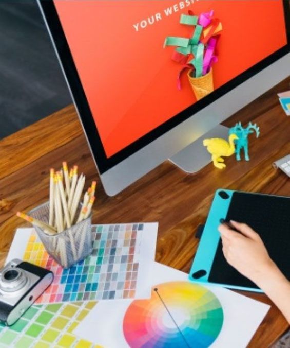 Workspace with computer displaying colorful design, a hand using a drawing tablet, color palettes, pencils, and a camera.