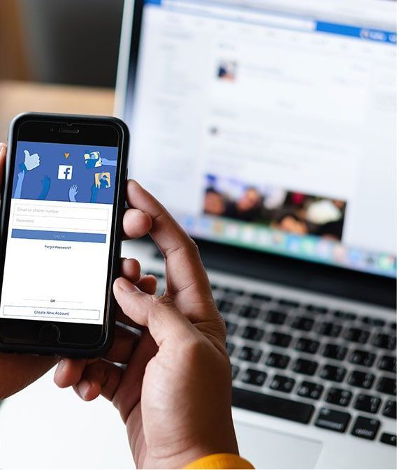 A person holding a smartphone with the Facebook login screen open, with a laptop displaying Facebook in the background.