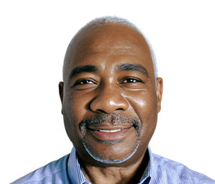 Smiling Black man with a shaved head and a graying goatee, wearing a blue collared shirt, against a white background.