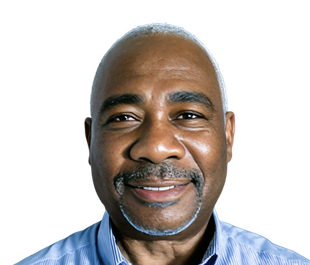Smiling Black man with a shaved head and a graying goatee, wearing a blue collared shirt, against a white background.