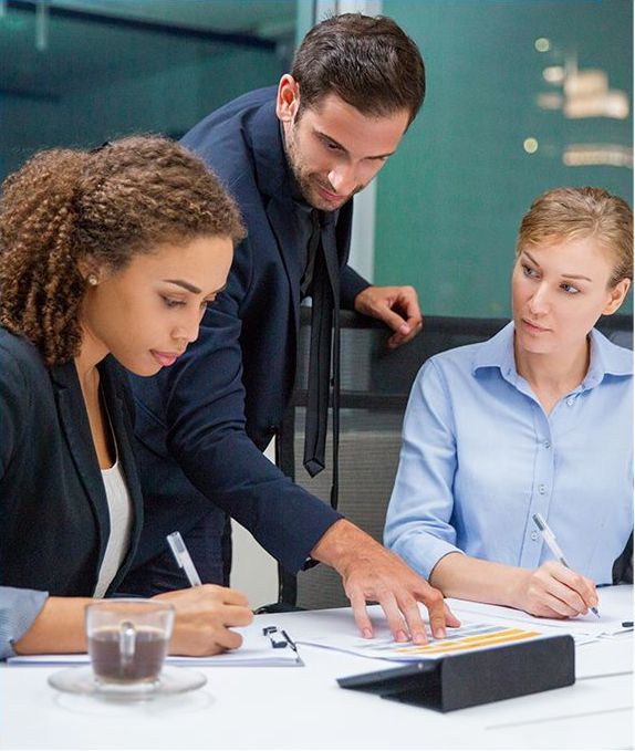 Three business professionals review documents at a table. A man in a suit points, while the women write and appear focused.