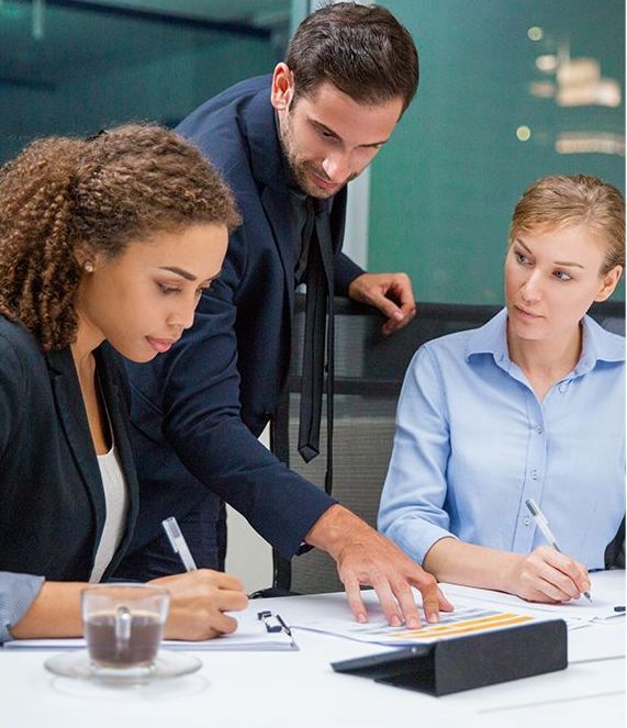Three business colleagues review documents at a table. A man points, while two women write.