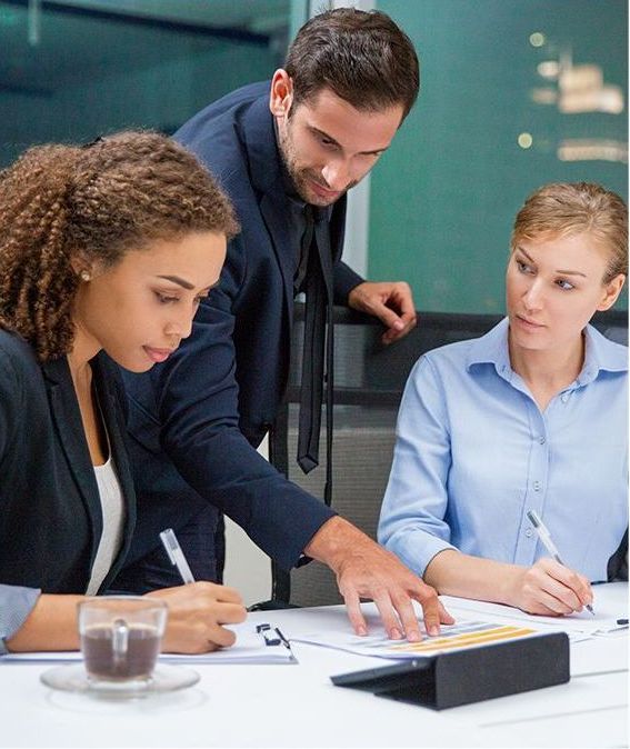 Three professionals in a modern office: a man pointing at documents, while two women write; focused expressions.