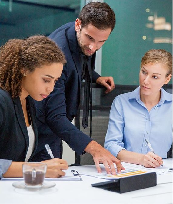 Three professionals in a meeting, a man pointing to a document while two women write. They are in an office setting, looking at the document.