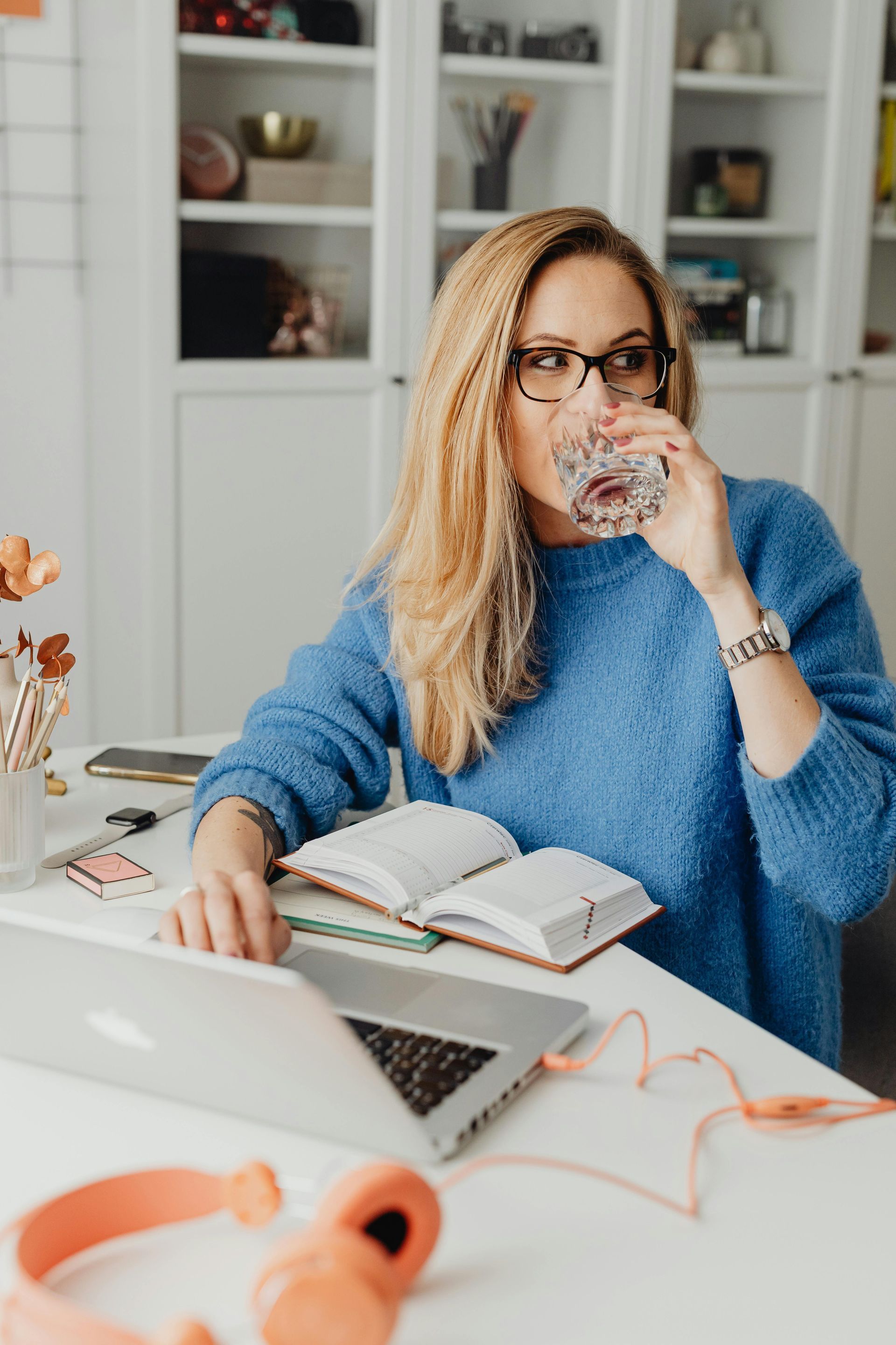 Blonde woman in blue sweater drinks from a glass while working on a laptop, wearing glasses.