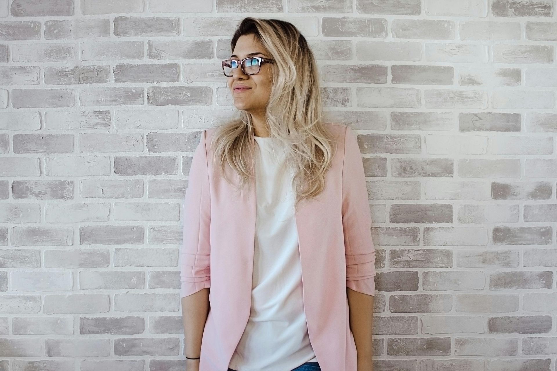 Woman with glasses and pink blazer leans against a light brick wall.