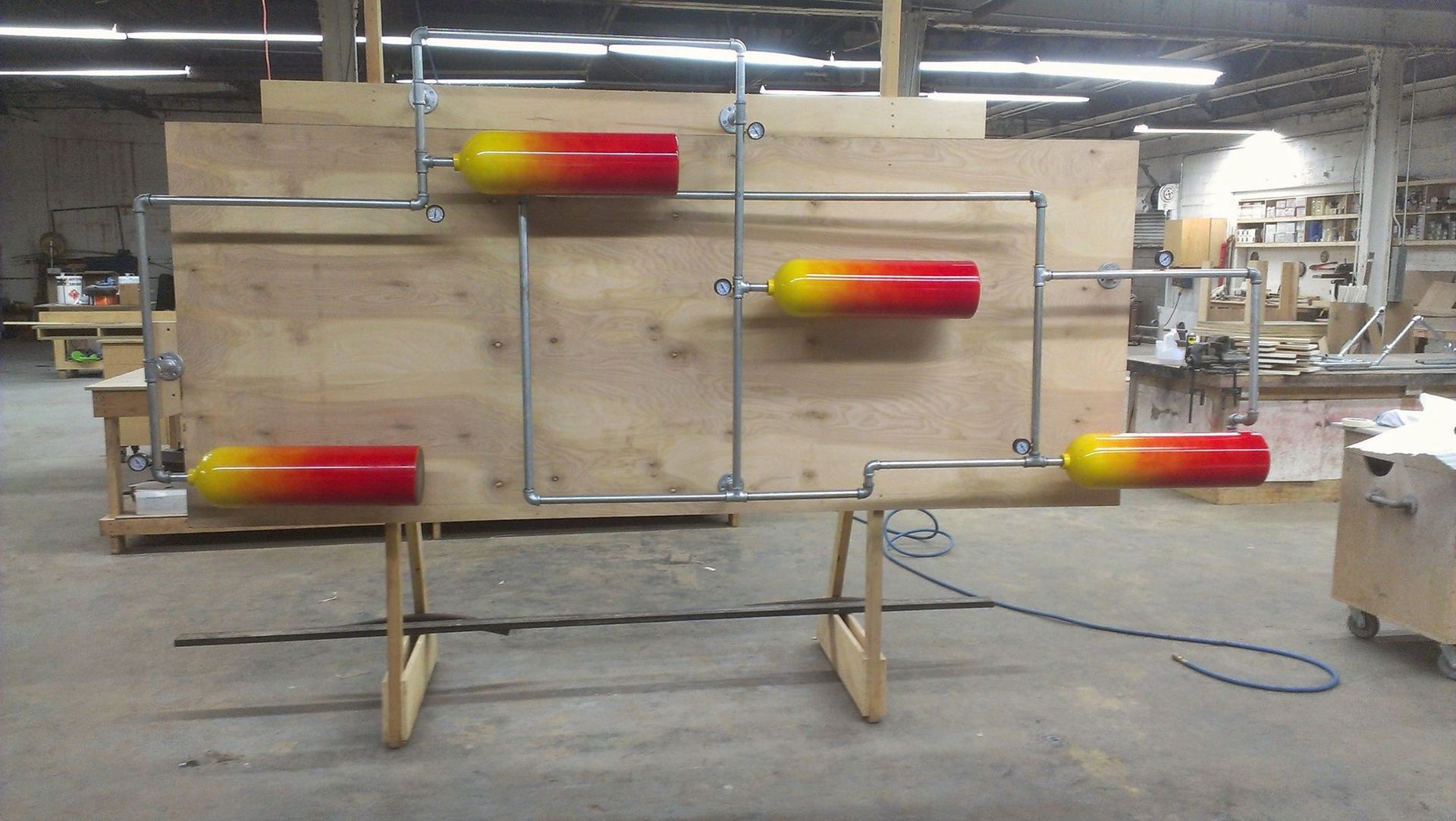 A wooden display with colorful cylinders connected by silver pipes, mounted on sawhorses in a workshop.