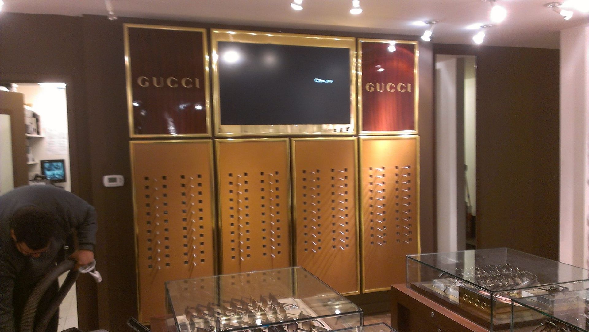 Gucci display unit with TV screen, gold accents, and watch display in a store. A person vacuums the floor.