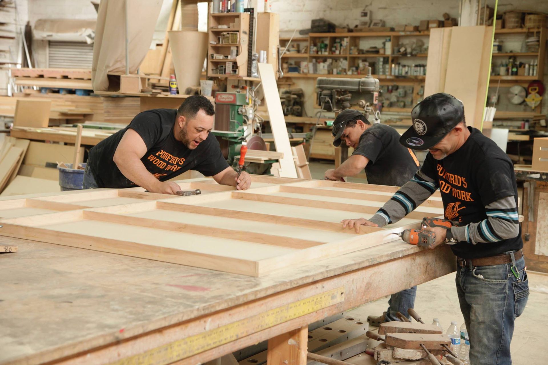 Three people in a woodshop building a wooden structure; one uses a drill.