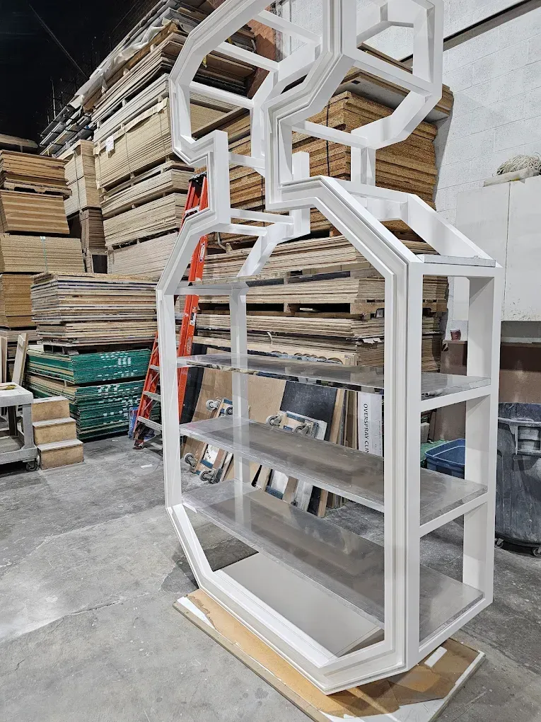 White geometric bookshelf structure in a wood shop with lumber in the background.