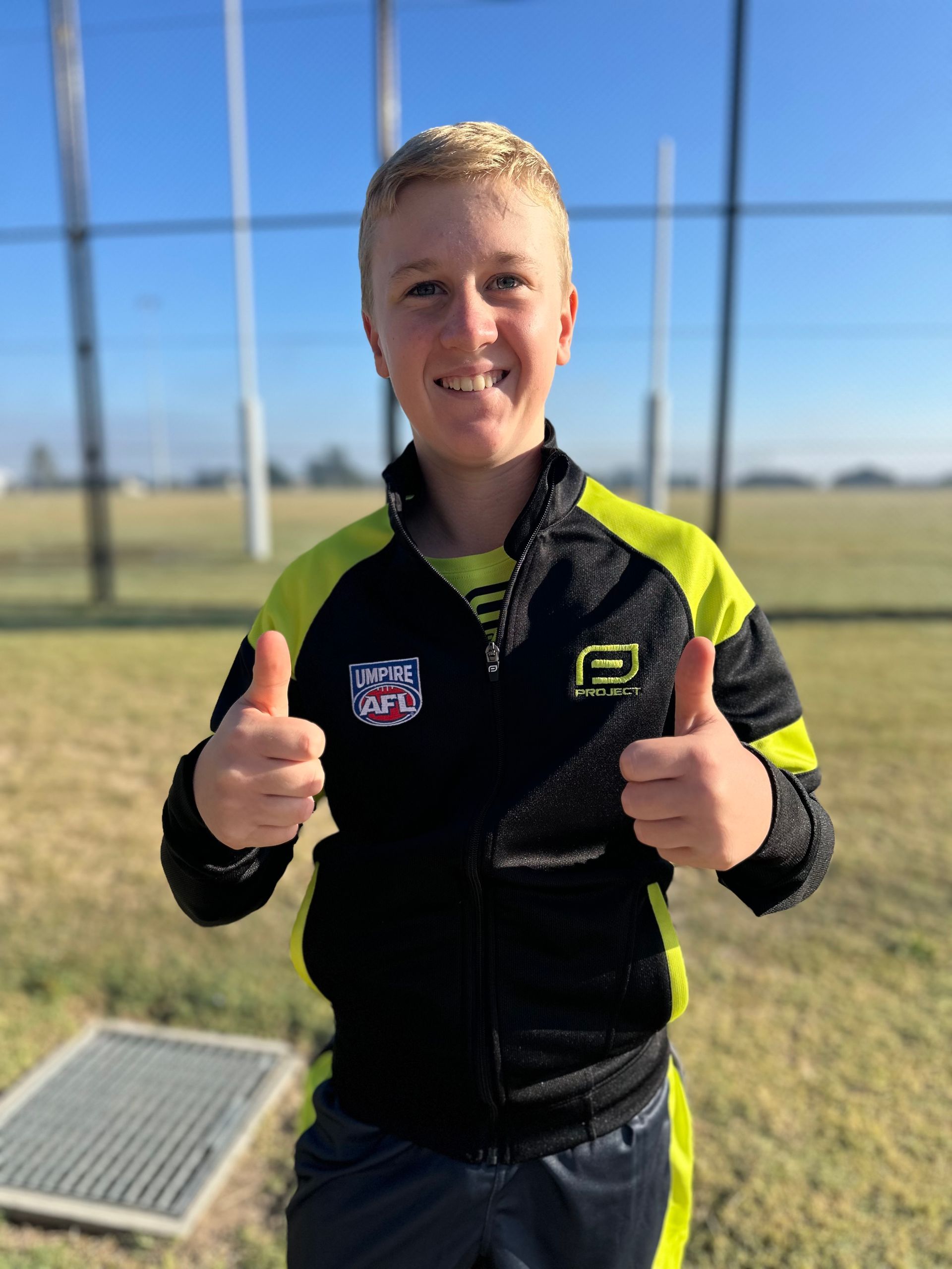 Young person in black and yellow jacket gives thumbs up on a sports field.