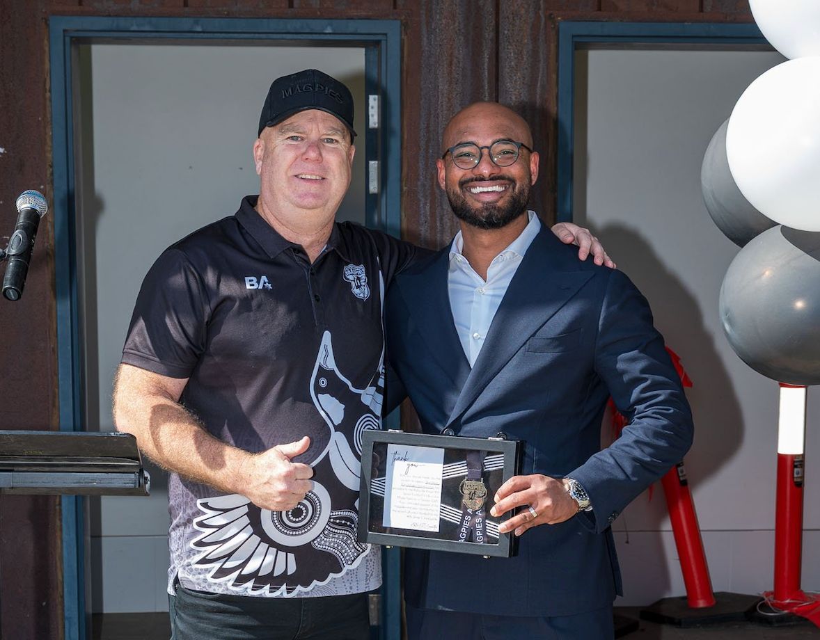 Two men, one in a black cap and shirt, the other in a suit, pose with a plaque.