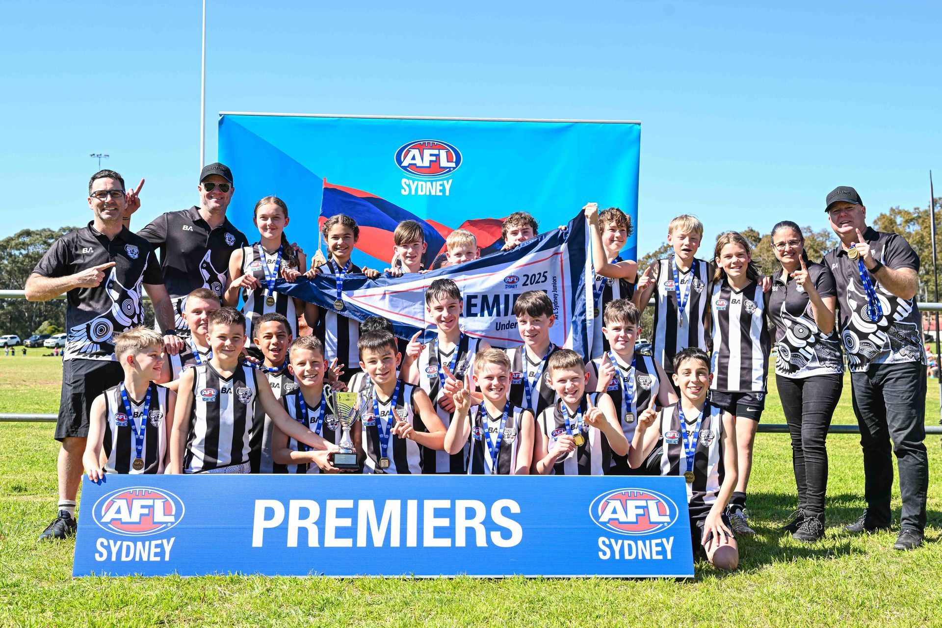 The 2025 Under 12 Div 2 team celebrates a premiership win outdoors, holding a banner and trophy.