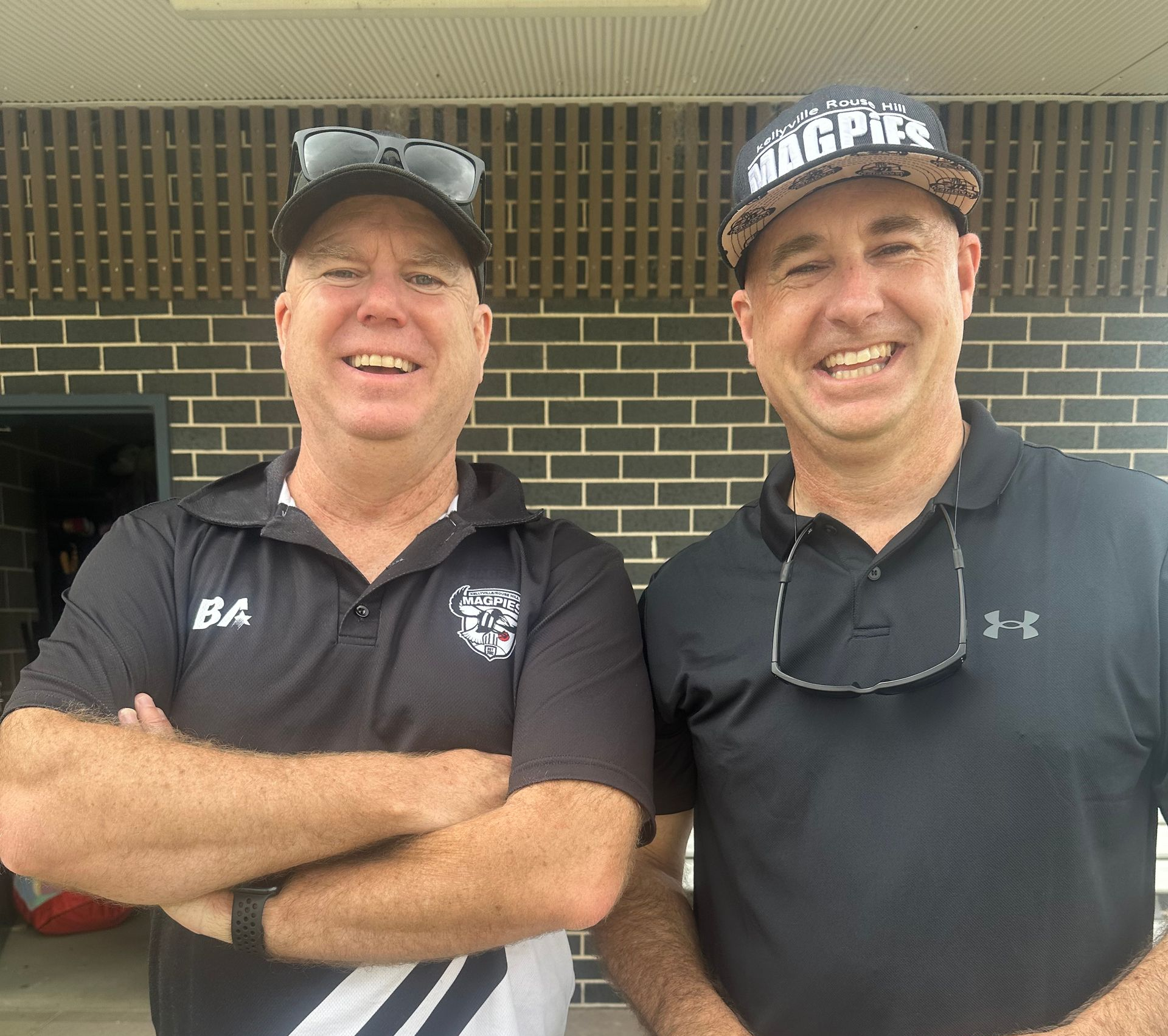 Two men smiling, one with arms crossed, in front of a brick wall. One wears a