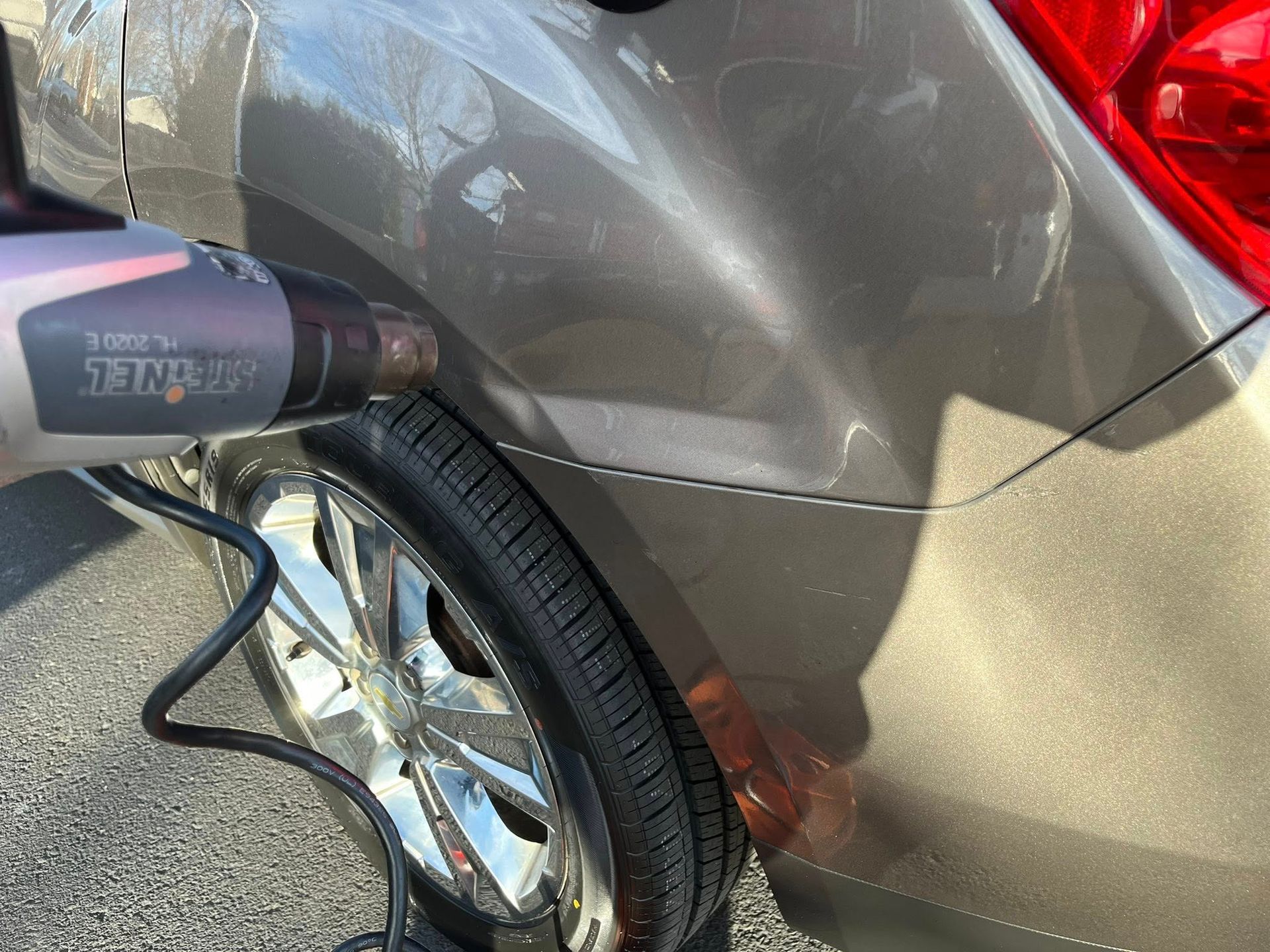 A heat gun is being held against a dent on the rear quarter panel of a gray car to assist in removing the damage.
