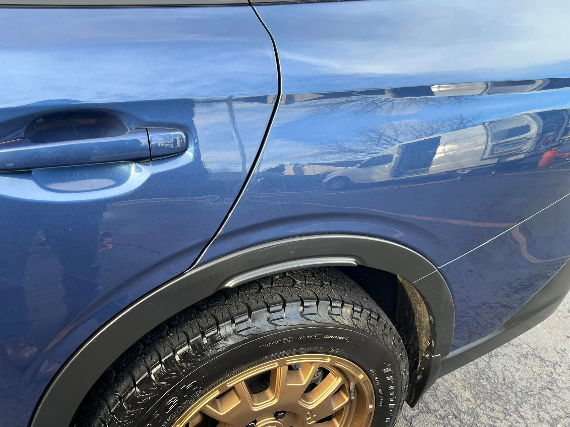 A side view of a blue car's rear wheel well, featuring a black trim piece, a bronze wheel, and a tire with tread.
