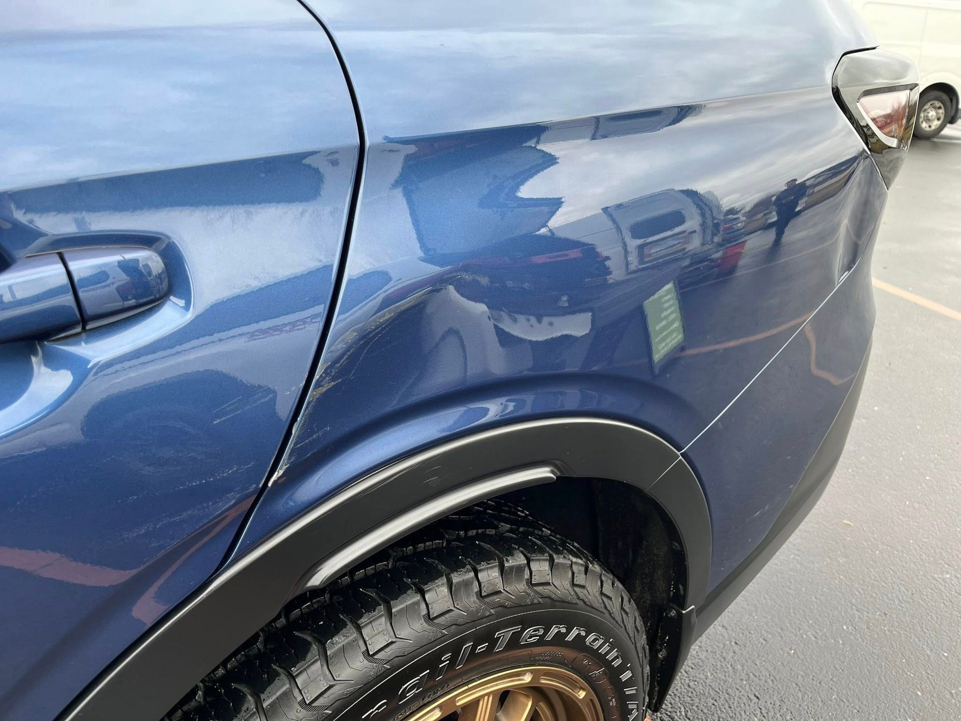 A close-up view of the rear quarter panel of a blue SUV, showing a dent above the black wheel arch trim.