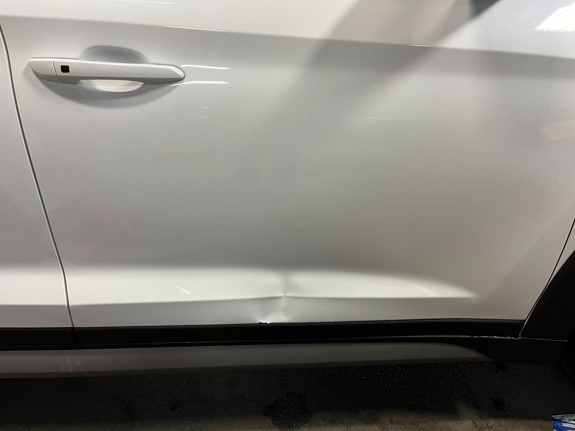 A side view of a white car door showing a dent on the lower body line near the bottom trim.