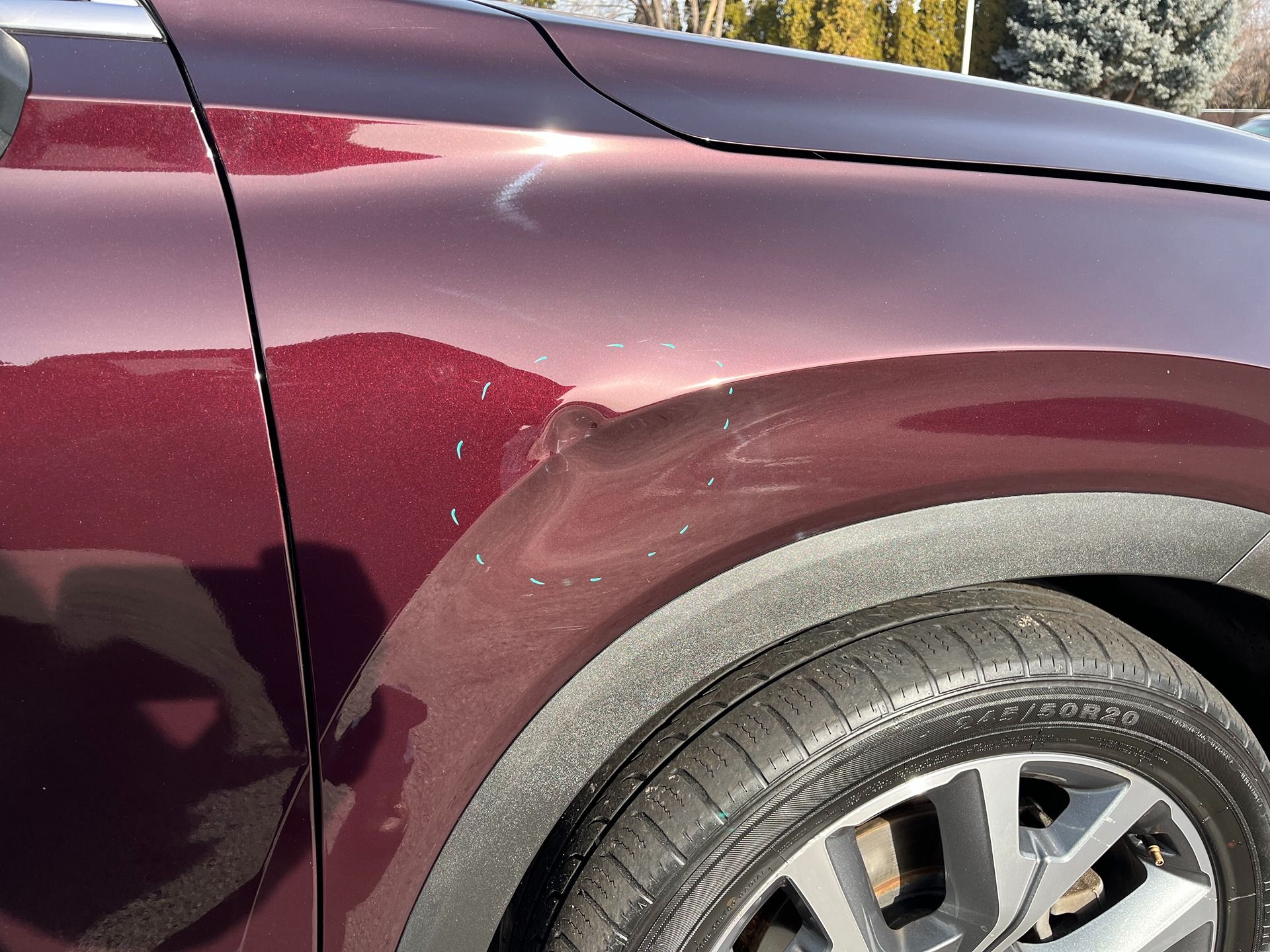 A close-up view of a maroon car's front fender showing a dent and surface scratches above the wheel well.