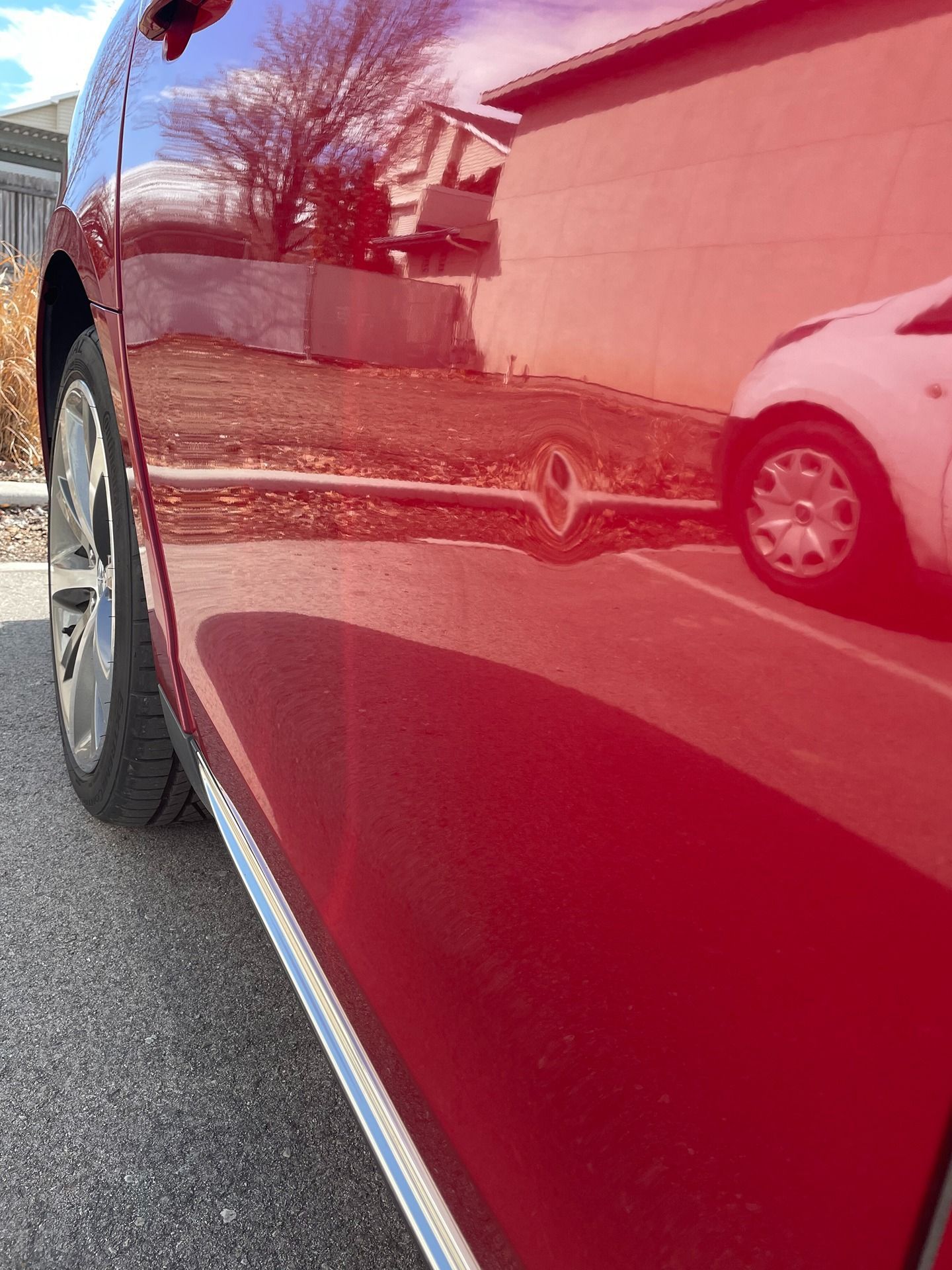 A close-up view of the side of a shiny red car, showing a small dent reflecting a nearby white vehicle and building.