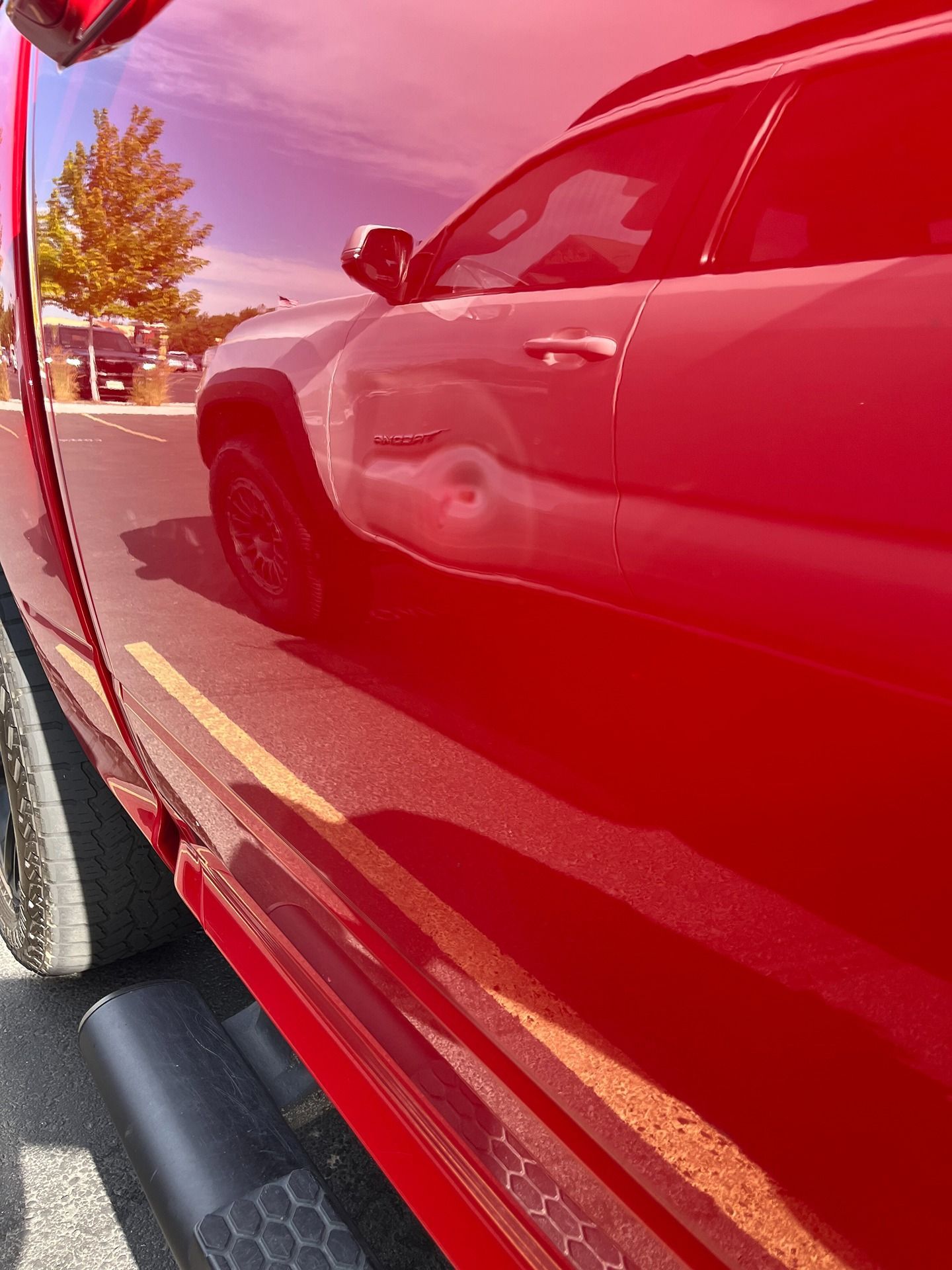 Close-up of a bright red truck's side panel, reflecting a parked car and trees on its glossy surface.