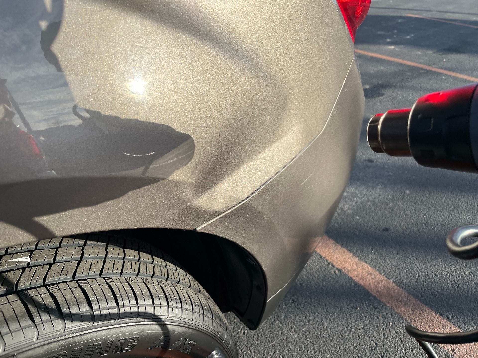 A heat gun pointed at a shallow dent on the bronze-colored rear quarter panel of a vehicle parked on asphalt.