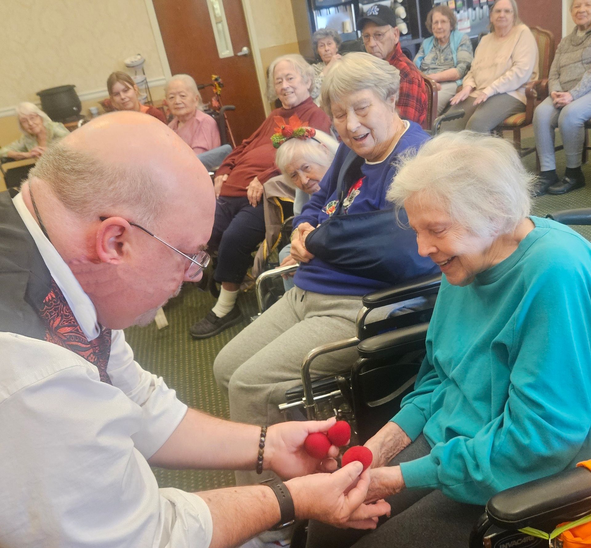 A person performs a magic trick with red foam balls for a group of seated residents in a community room.