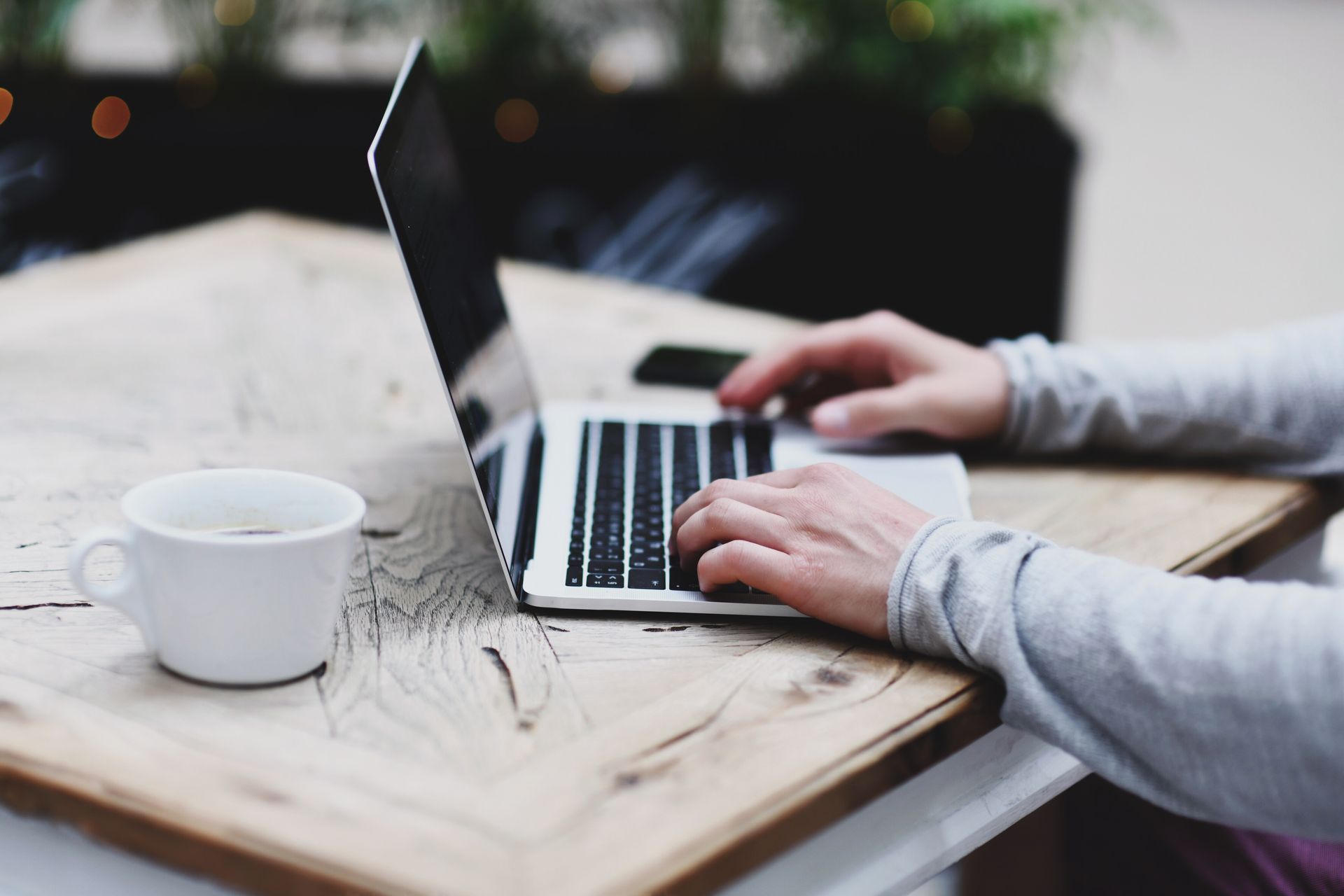 Eine Person tippt an einem Laptop an einem Holztisch mit einer Kaffeetasse in der Hand.