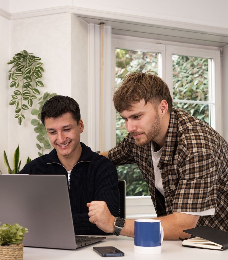 Zwei Männer sitzen an einem Tisch und schauen auf einen Laptop.