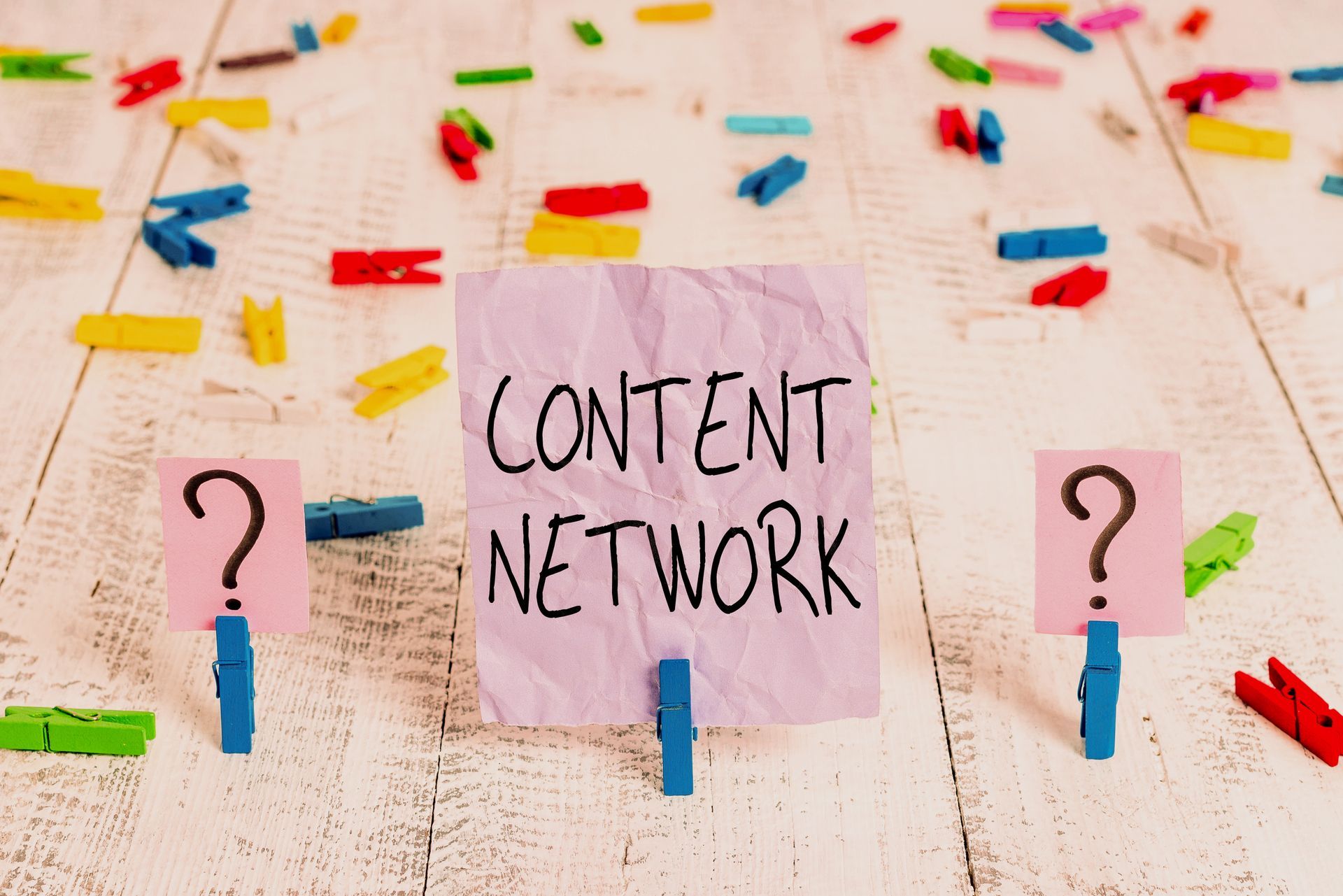 Rosa Zettel mit der Aufschrift „CONTENT NETWORK” und Fragezeichen auf Wäscheklammern, bunte Klammern verstreut auf Holz.