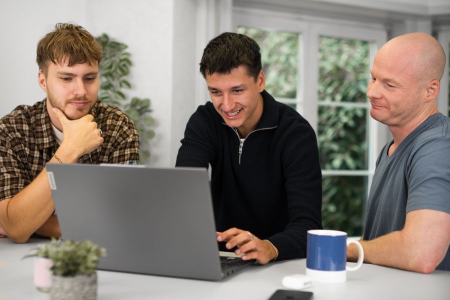 Drei Männer sitzen an einem Tisch und schauen auf einen Laptop.