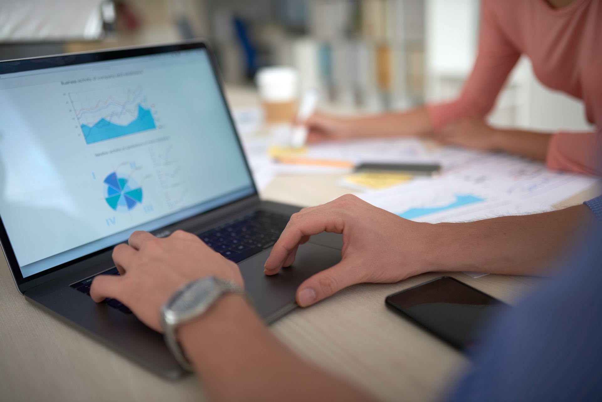 Person, die einen Laptop benutzt, wahrscheinlich Daten analysiert, mit einer Uhr, neben einem Telefon. Eine weitere Person ist im Hintergrund zu sehen.