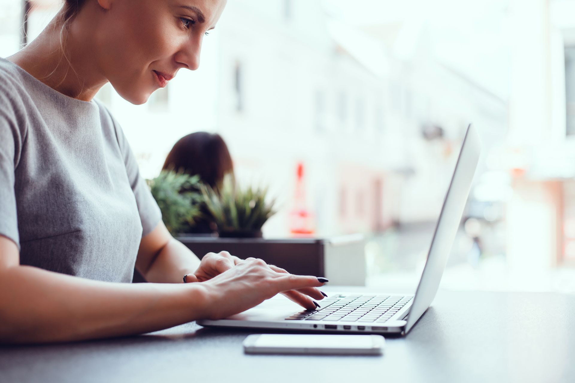 Frau, die an einem Tisch in der Nähe eines Fensters einen Laptop benutzt; daneben liegt ein Smartphone.