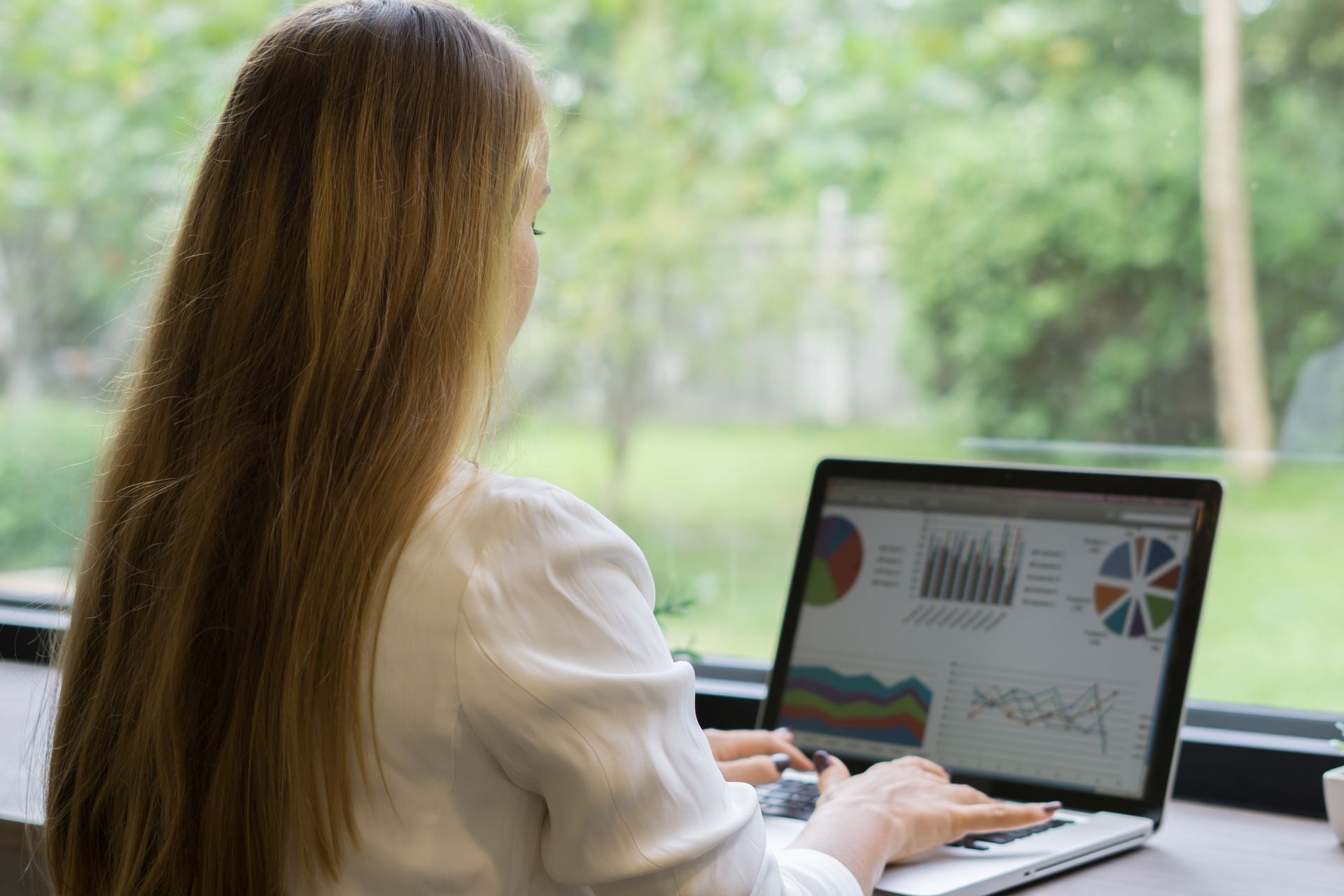 Frau mit langen blonden Haaren, die an einem Laptop arbeitet und Diagramme anzeigt, in der Nähe eines Fensters mit Bäumen.