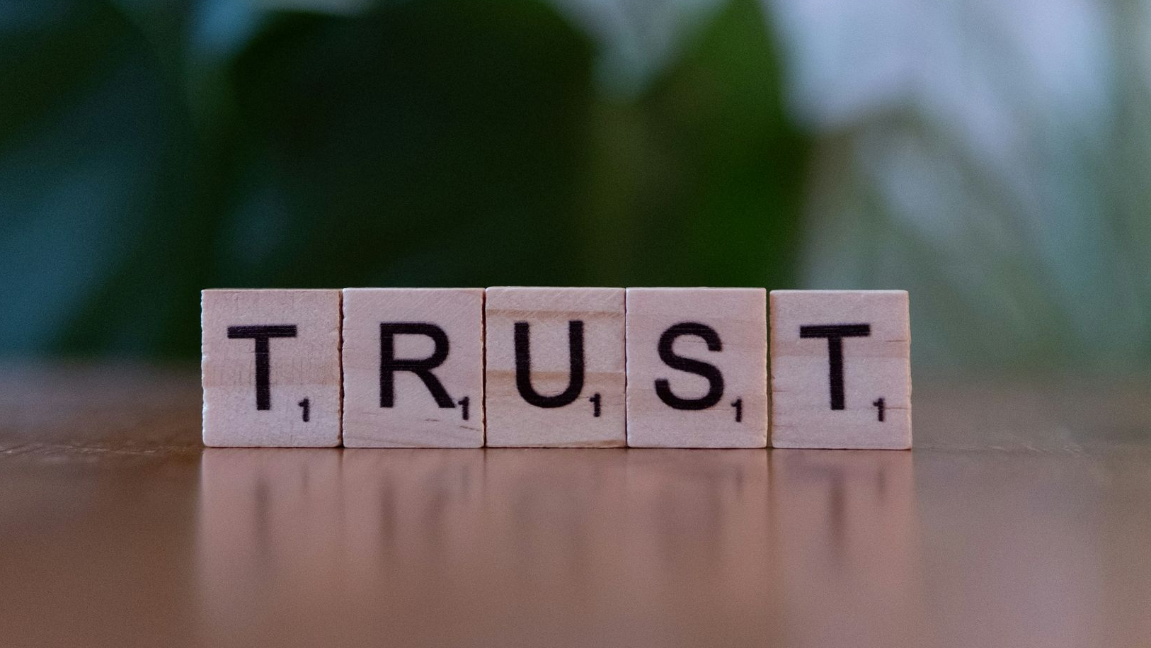„TRUST“ aus Scrabble-Steinen auf einer Holzoberfläche, mit verschwommenem grünen Hintergrund.