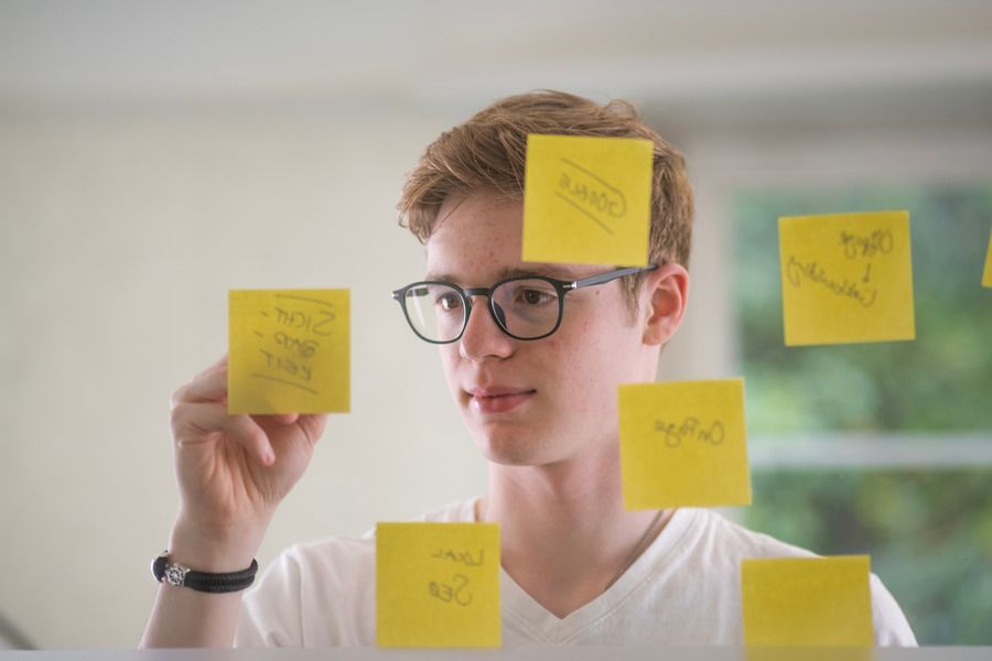 Ein junger Mann mit Brille betrachtet Haftnotizen an einer Glaswand.