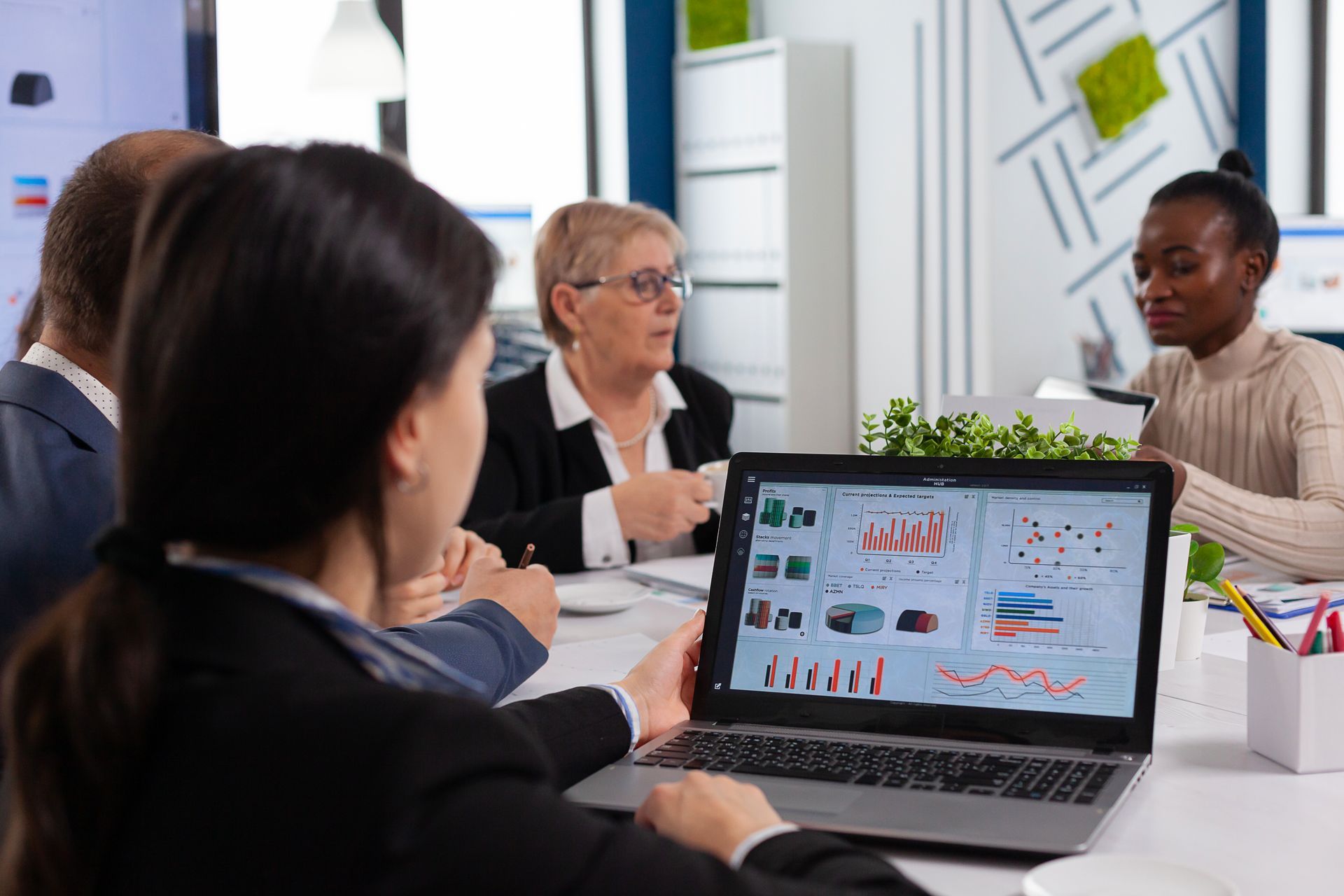 Mehrere Personen sitzen an einem Tisch und führen eine Besprechung; eine Frau blickt dabei auf ein Laptop mit Diagrammen und Analysen.