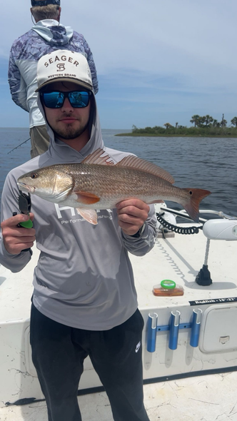 A person in a gray hooded shirt and hat holds a redfish on a boat, with another person in the background.